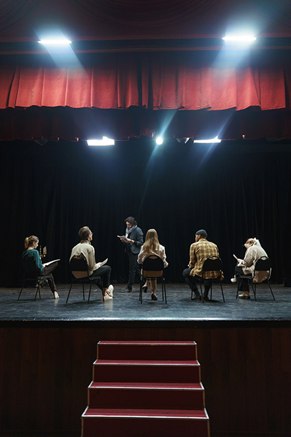 Eine Theaterbühne mit Treppe und sitzende Darsteller sowie stehender Dozent